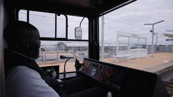 Interior shot of a MetroLink operator driving a train by the Lambert Airport Terminal #2 station. The platform of the station can be seen through the windshield.