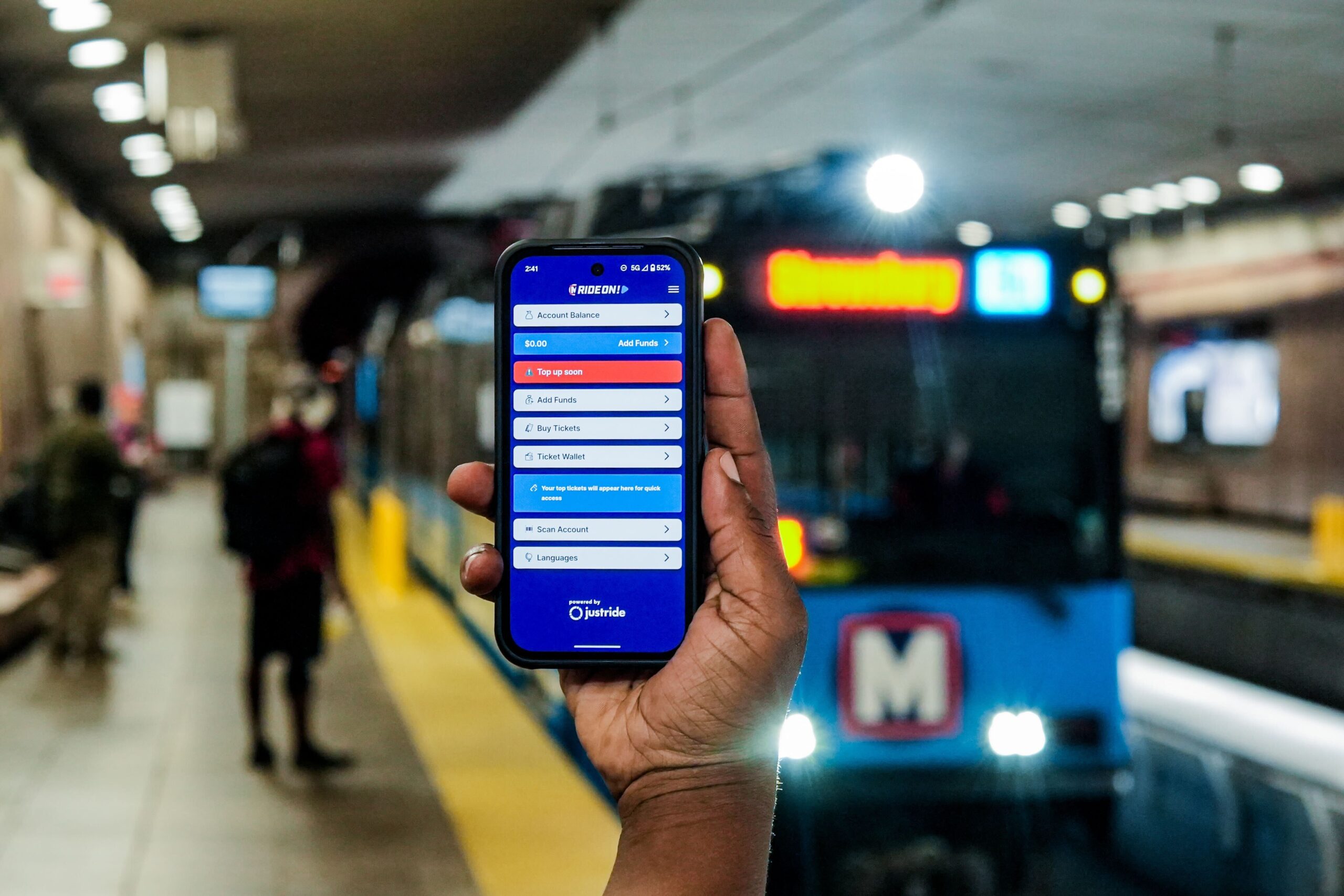 A hand holds a phone with Metro's RideOn app while a MetroLink train pulls into a station in the background.